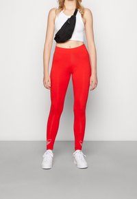 Rode leggings met een gladde textuur, hoge tailleband, met een wit ontwerpelement bij de enkel. Gecombineerd met een cropped wit topje en een zwarte schoudertas.