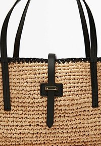 Sac cabas tressé en paille avec des accents et des poignées en cuir noir. Il présente une sangle plate noire et un détail rectangulaire en métal.