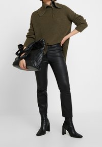 Maglione a maglia verde oliva con colletto alto, pantaloni in finta pelle neri e una borsa a tracolla nera intrecciata con spalline spesse e base strutturata.