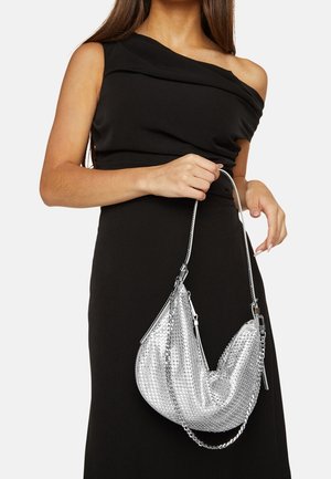 Nainen mustassa olkaimeton mekossa, joka pitää kiiltävää hopeanväristä, kuvioitua käsilaukkua, jossa on ohut hihna, valkoista taustaa vasten.