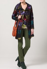 Cappotto multicolore e fantasia con un grande colletto, indossato sopra una blusa floreale. Abbinato a pantaloni verde oliva e scarpe con tacco verde scuro.