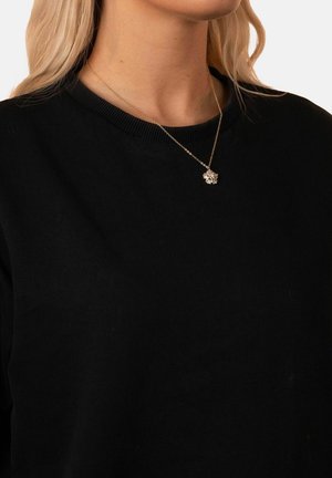 Blonde Frau, die einen schwarzen Pullover und eine goldene Halskette mit einem detaillierten blumenförmigen Anhänger trägt.