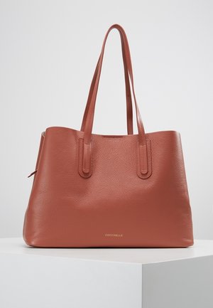 Sac fourre-tout en cuir texturé marron avec deux longues poignées, logo discret près du centre inférieur, présenté sur un socle blanc contre un fond uni.