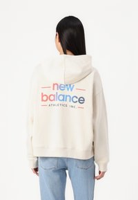 Felpa color crema con cappuccio regolabile. Caratterizzata da un logo "new balance" colorato e il testo "ATHLETICS INC." sul retro. Vestibilità ampia.
