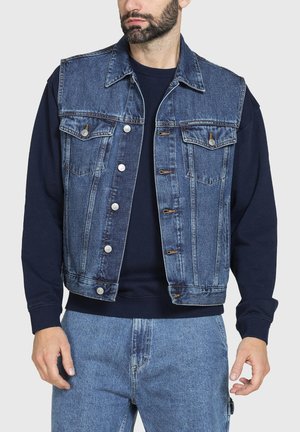 Uomo che indossa un gilet denim lavaggio medio con bottoni argento sopra una felpa blu navy e jeans azzurri, in piedi su uno sfondo semplice.