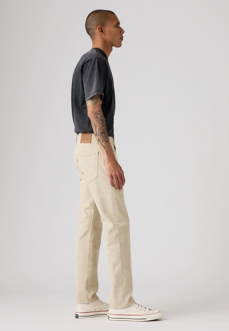 Uomo con braccio tatuato che indossa una maglietta nera, jeans beige e scarpe da ginnastica bianche, in piedi di lato contro uno sfondo chiaro uniforme.