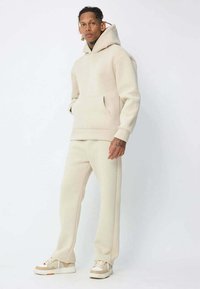 Sweat-shirt à capuche beige avec une poche frontale et un pantalon de survêtement assorti. Le matériau a une texture douce ; les caractéristiques incluent des poignets et une taille élastiques.