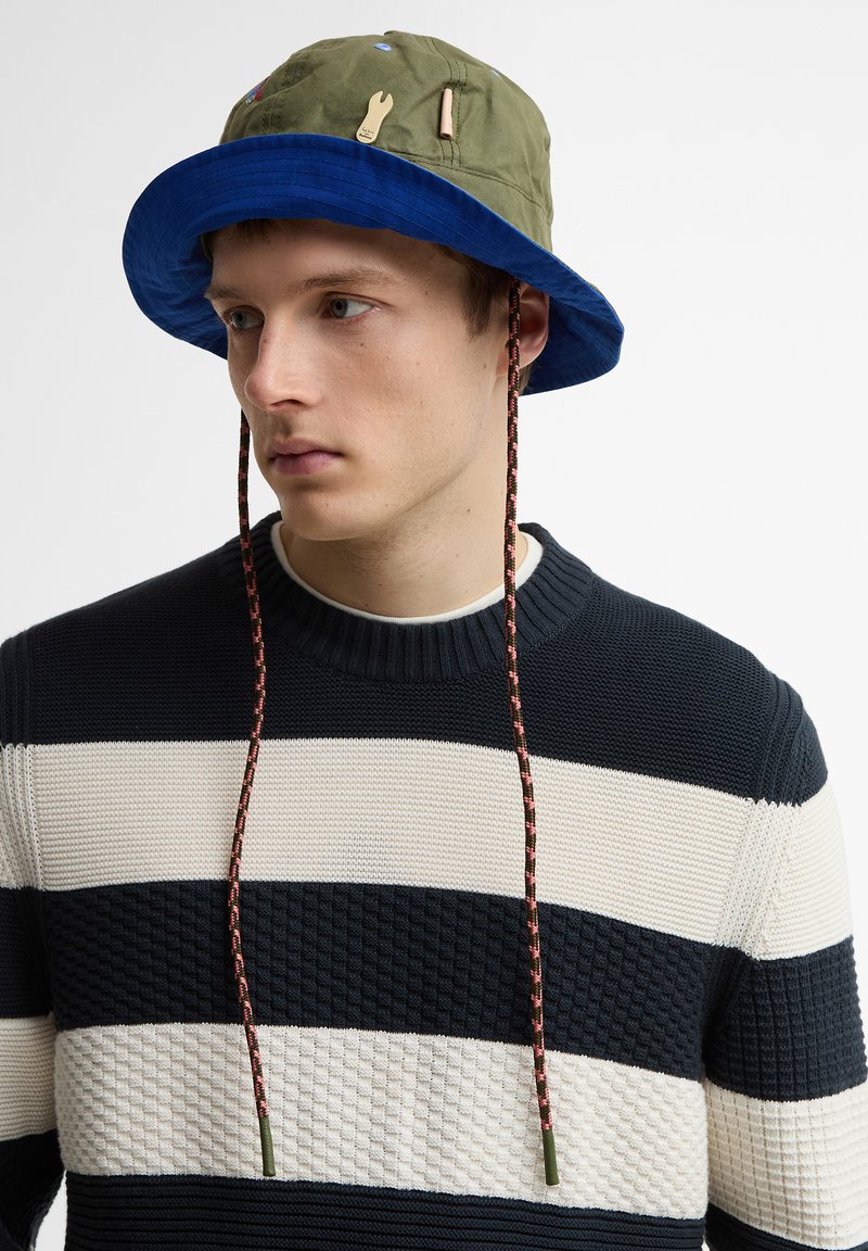 Νεαρός άνδρας που φοράει καπέλο τύπου bucket σε ελιά και μπλε χρώμα με λουράκια στο πιγούνι, ντυμένος με ριγέ μαύρο και άσπρο πλεκτό πουλόβερ, κοιτάζοντας προς τα αριστερά.