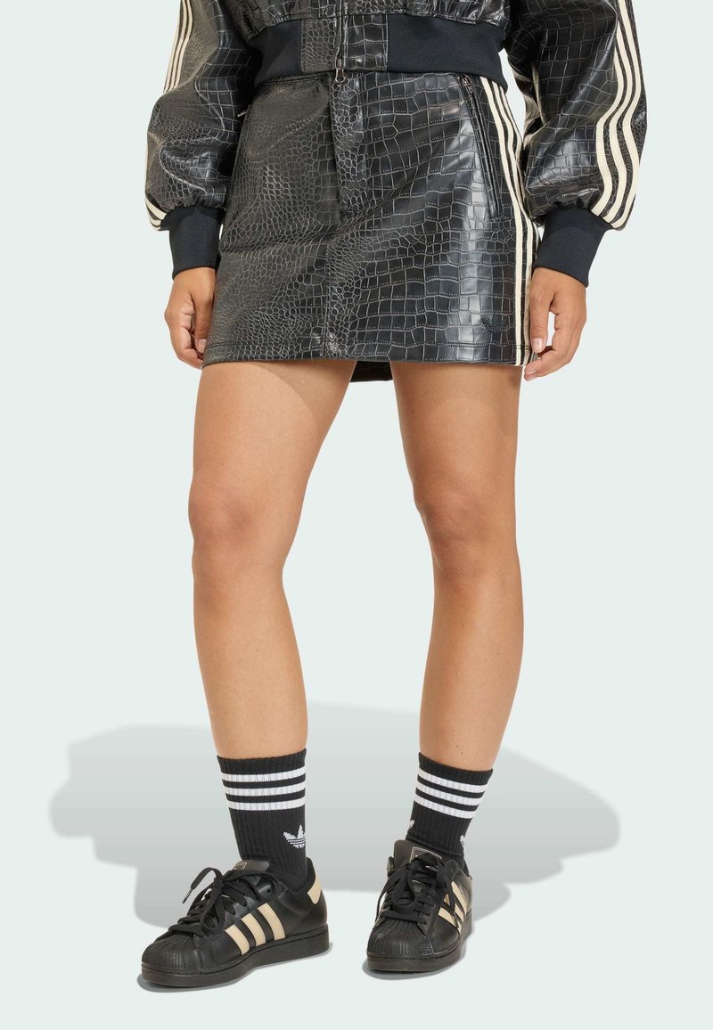 Černá lesklá minisukně se vzorem krokodýlí kůže, doplněná černými teniskami Adidas s bílými pruhy a černobílými sportovními ponožkami.