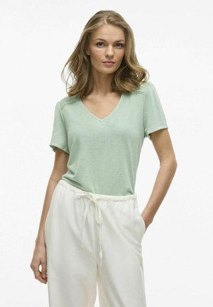 Camiseta de algodón verde claro con escote en V y mangas cortas, con detalles de encaje en los hombros, combinada con pantalones blancos de cintura elástica.