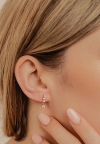 Goudkleurige oorbellen met drie kleine heldere edelstenen in een verticale lijn, genesteld tegen een geperforeerd oorlel. Gladde huid en naakte nagels zichtbaar.
