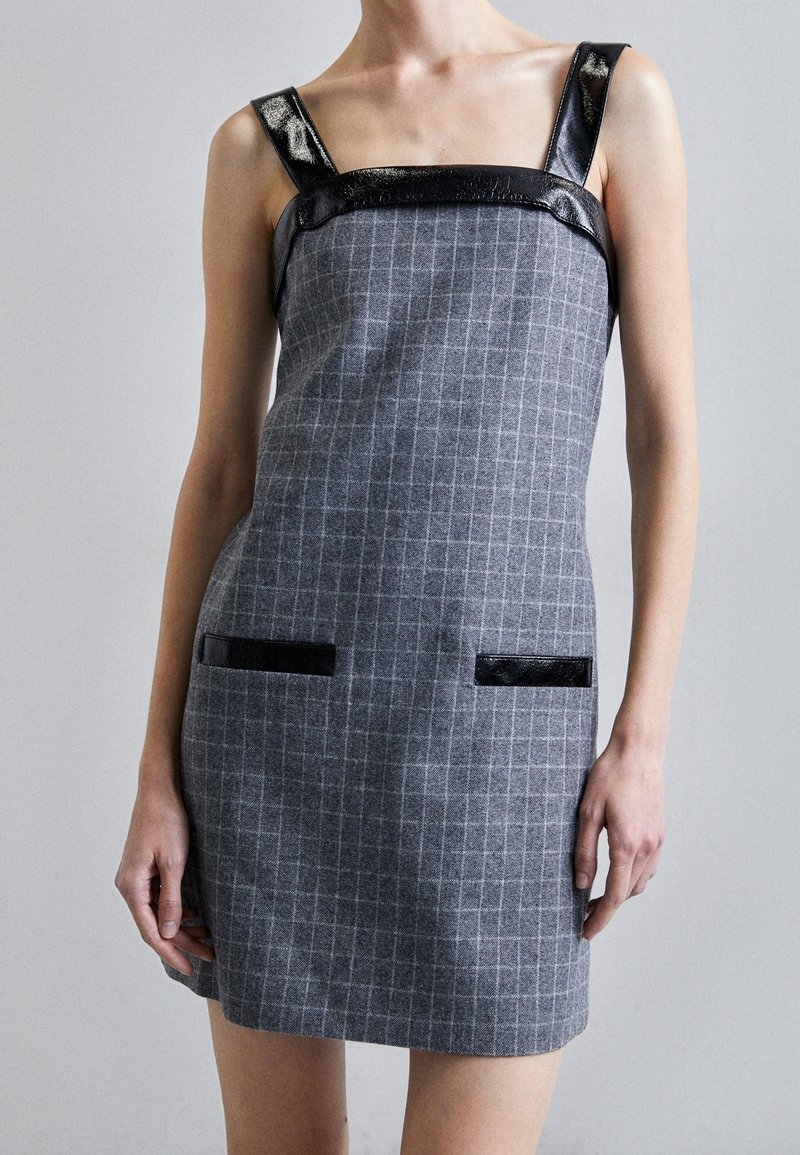 Robe à carreaux grise sans manches avec bretelles en cuir noir et bordure portée par une personne, montrée du cou à la cuisse sur un fond uni.
