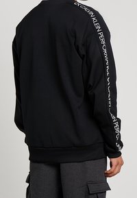 Sweat-shirt noir à manches longues avec un logo blanc le long de la manche gauche, poignets côtelés et coupe décontractée. Texture lisse.