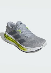 Zapatilla de running gris con parte superior de malla, tres rayas negras, detalles reflectantes, adornos en verde lima en la suela y una entresuela blanca acolchada.
