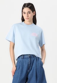 Ljust blå bomull t-shirt med korta ärmar och rundad halsringning, med en rosa grafisk logotyp på vänster sida. Avslappnad passform.