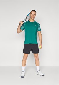 Camiseta atlética verde con mangas estampadas, pantalones cortos negros, calcetines blancos y zapatillas deportivas blancas. Sosteniendo una raqueta de tenis y una pelota.