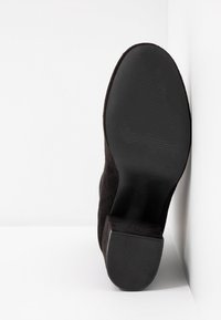 Botte en daim noire reposant semelle vers le haut contre un coin blanc, montrant une semelle en caoutchouc lisse et un talon bloc avec texture antidérapante.