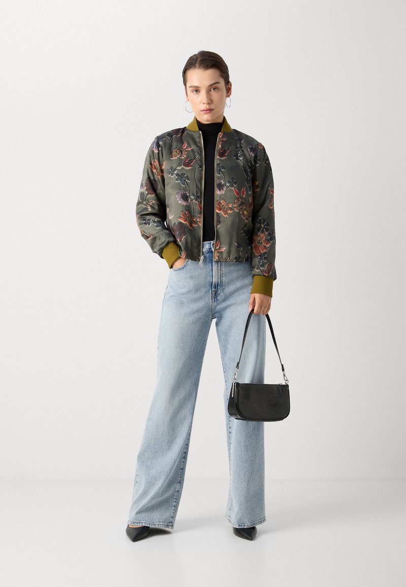 Veste bomber à motif floral en satin vert foncé, avec des poignets côtelés olive, assortie à un jean bleu clair taille haute et un petit sac à main noir.