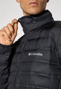 Vyras, vilkintis juodą įsagų Columbia striukę, traukia užtrauktuką prie apykaklės, matomas logotipas ant krūtinės.