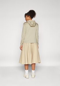 Kapucnis, világos bézs felső, laza szabással, egy áramló bézs szoknyával párosítva. A modellen fehér bokazokni és világos sportcipő van.