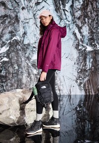 Dark purple waterproof jacket, black backpack with logo, black boots with white soles, leggings, light gray socks, and pink cap.