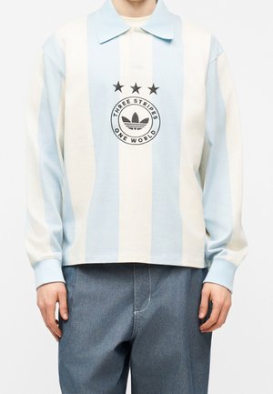 Persona que lleva una camiseta tipo polo de manga larga, con rayas verticales en azul claro y blanco, con un logo de "Three Stripes One World" y tres estrellas encima.