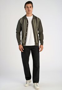Olijfgroen cardigan met verticale witte strepen, voorzien van een knopenrij aan de voorkant en een gespreide kraag, gedragen over een wit T-shirt en zwarte jeans.
