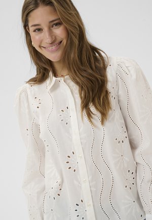 Balta izšūta blūze ar ziedu izgriezumiem, pogām priekšpusē un pūkainām piedurknēm. Smalka tekstūra ar viļņotu rakstu visā blūzē.