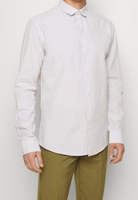 Camicia a maniche lunghe grigio chiaro con bottoni e colletto, caratterizzata da una texture liscia e un orlo arrotondato, indossata con pantaloni verde oliva.