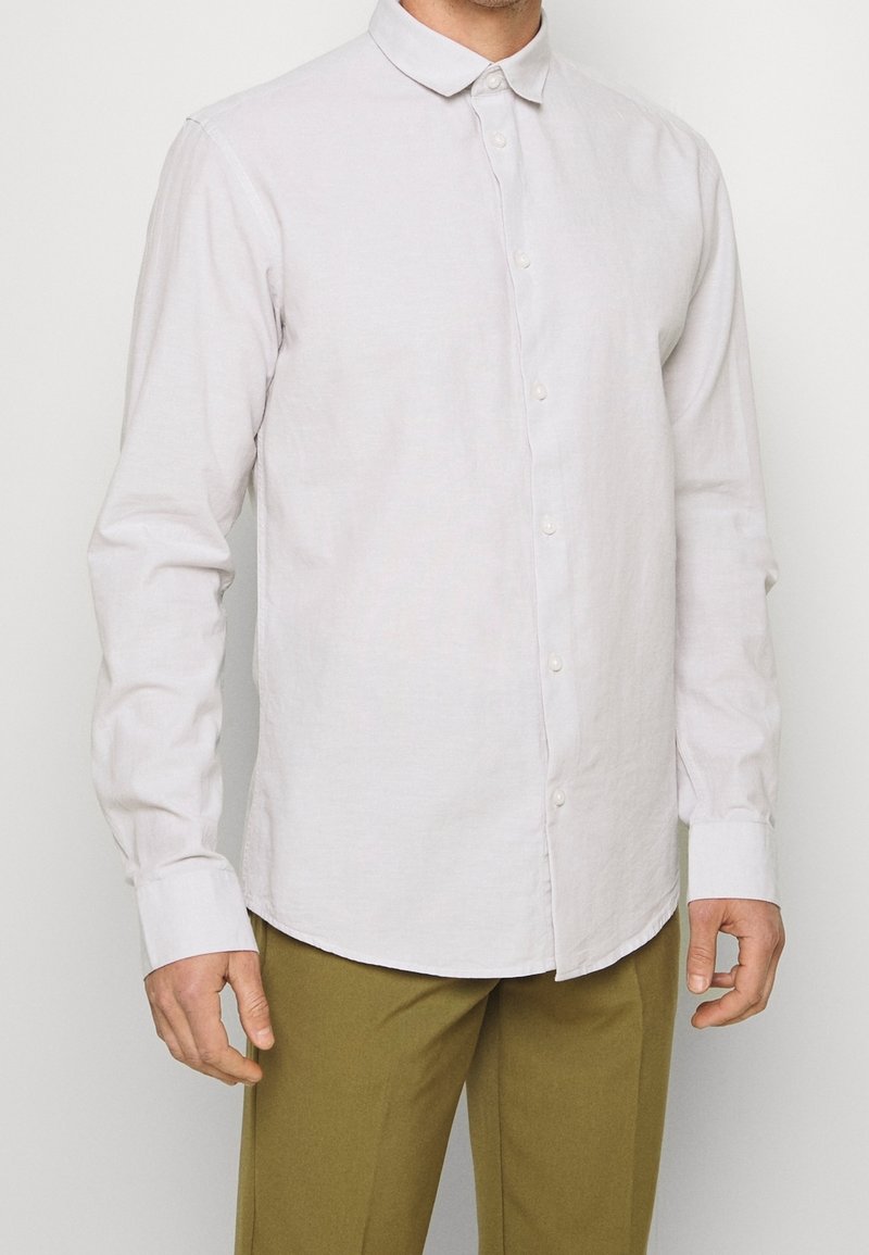 Camicia a maniche lunghe grigio chiaro con bottoni e colletto, caratterizzata da una texture liscia e un orlo arrotondato, indossata con pantaloni verde oliva.