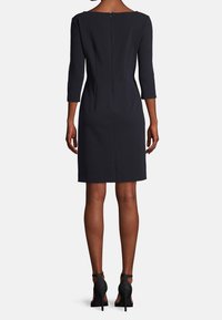 Abito navy con maniche a tre quarti, forma aderente e dettaglio della zip posteriore. Tessuto testurizzato, lunghezza al ginocchio, abbinato a scarpe con tacco alto nere.
