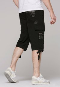Černé cargo šortky s několika kapsami a nastavitelnými stahovacími šňůrkami na lemu. V kombinaci se světle šedými sportovními botami, umístěné na neutrálním pozadí.