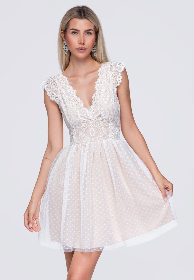 Femme blonde portant une robe en dentelle blanche avec un décolleté en V profond et une jupe à pois, tenant l'ourlet d'une main sur un fond uni.