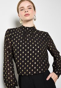 Schwarze Bluse mit goldenem Ovalmuster, hochgeschlossen, lange Ärmel mit Raffung, aus leichtem Stoff gefertigt. Akzentuiert mit goldenen Ohrringen.