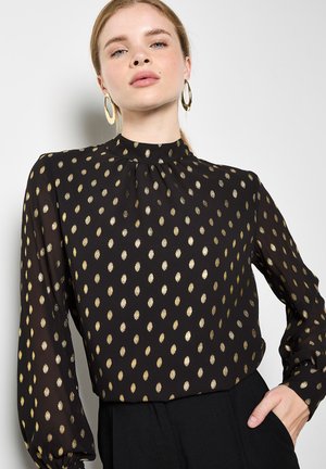 Blusa negra con un patrón de óvalos dorados, cuello alto, mangas largas con detalle fruncido, hecha de un tejido ligero. Acentuada con pendientes dorados.