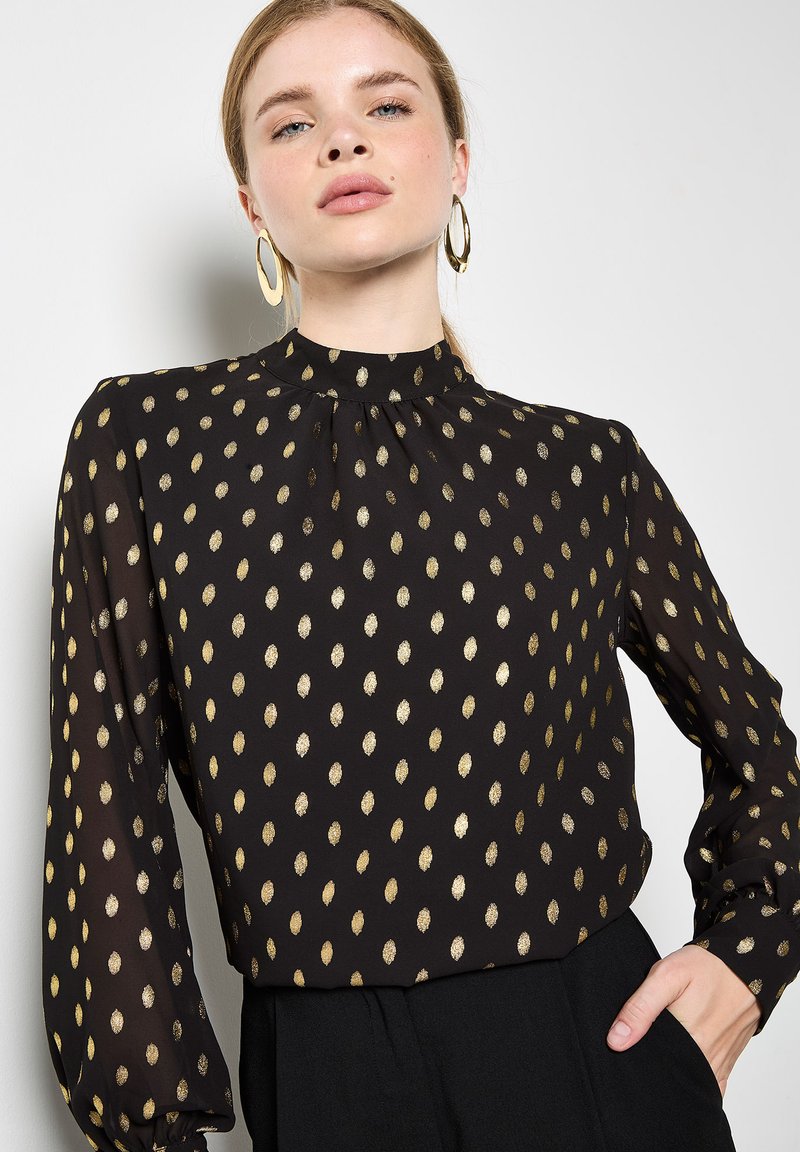 Schwarze Bluse mit goldenem Ovalmuster, hochgeschlossen, lange Ärmel mit Raffung, aus leichtem Stoff gefertigt. Akzentuiert mit goldenen Ohrringen.