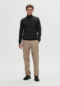 Mladić stoji odjeven u crnu duksericu sa dugim rukavima i ovratnikom na zip, bež hlače i crne cipele protiv svjetlosti na jednostavnom svijetlom pozadini.