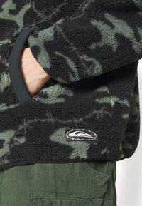 Schwarz-grüner Fleecepullover mit Tarnmuster, glatten Abschlüssen, seitlichen Taschen und einem Logo-Patch mit Stacheldraht-Design.