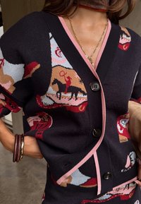 Γυναίκα με μαύρη ζακέτα με κουμπιά, ροζ διακόσμηση και πολύχρωμα σχέδια αλόγου και αναβάτη, με αξεσουάρ ένα χρυσό κολιέ και ξύλινες βραχιόλες.