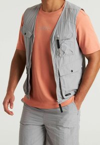 Gilet grigio chiaro senza maniche con più tasche; presenta un tessuto strutturato e una chiusura con cerniera frontale. Abbinato a una camicia corallo a maniche corte.