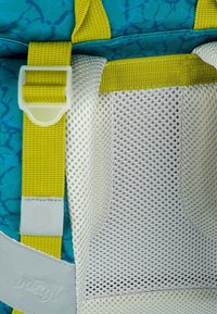 Rucksackdetails mit einer türkisfarbenen gemusterten Außenseite, gelbem Riemen, weißem Netzfach und einem grauen Etikett mit dezentem Branding.