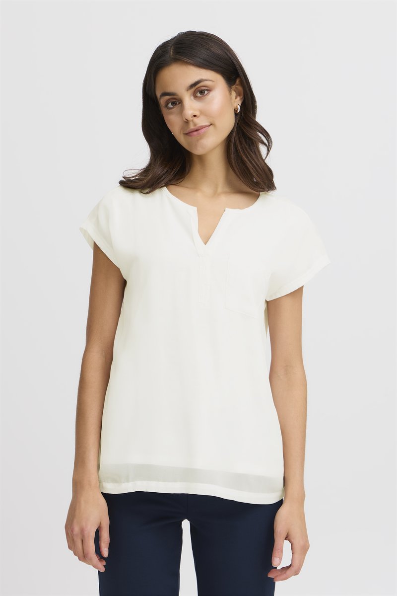 Jeune femme aux cheveux foncés portant un chemisier blanc à manches courtes et un pantalon foncé, debout devant un fond clair uni.