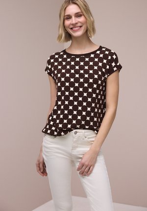 Uśmiechnięta kobieta o krótkich blond włosach, ubrana w czarny i biały top w groszki z krótkim rękawem i białe spodnie, stojąca na beżowym tle.