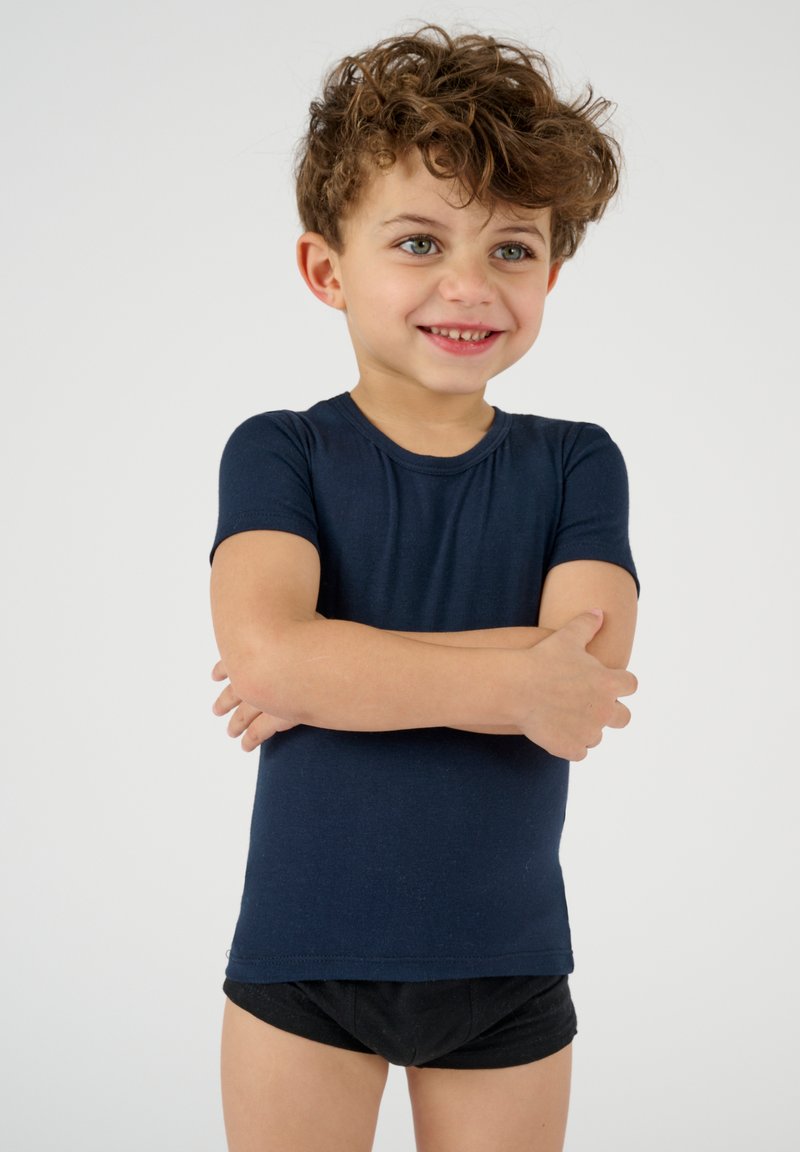 T-shirt fitted a maniche corte di colore blu navy, materiale morbido, colletto a girocollo, indossato con pantaloni corti neri, bambino in piedi con le braccia incrociate, sfondo chiaro.