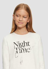 Weißes Langarmshirt mit einem schwarzen Textdesign, das "Nachtzeit" zeigt, ergänzt durch silberne und schwarze Perlenakzente. Aus weichem Baumwollmaterial gefertigt.