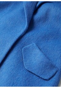 Un abrigo de fieltro azul con un cuello doblado y un pequeño bolsillo en forma de diamante. La textura es suave y ligeramente peluda.