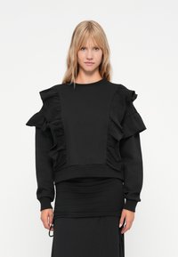 Hanorac negru cu accente volanate proeminente pe umeri �și un design ajustat. Fabricat dintr-un material moale cu o textură mată.