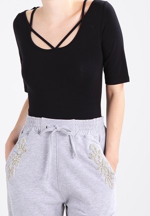 Frau trägt ein schwarzes Kurzarmtop mit überkreuzten Trägern und hellgraue Jogginghose mit dekorativer Stickerei auf den Taschen.