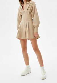 Beige klänningsskjorta med en figurnära överdel, långa puffärmar, plisserad kjol och knäppning framtill, i kombination med vita sneakers och ljusa strumpor.