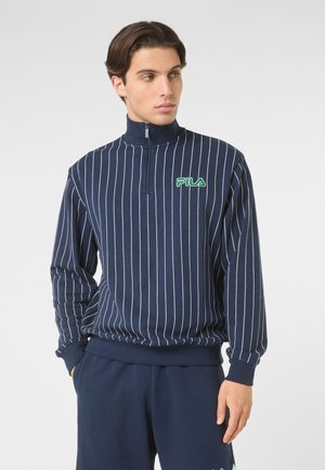 Mand iført en navyblå kvart-zip sweatshirt med tynde striber og FILA-logo, hænderne i lommerne, kigger til siden mod en ensfarvet baggrund.